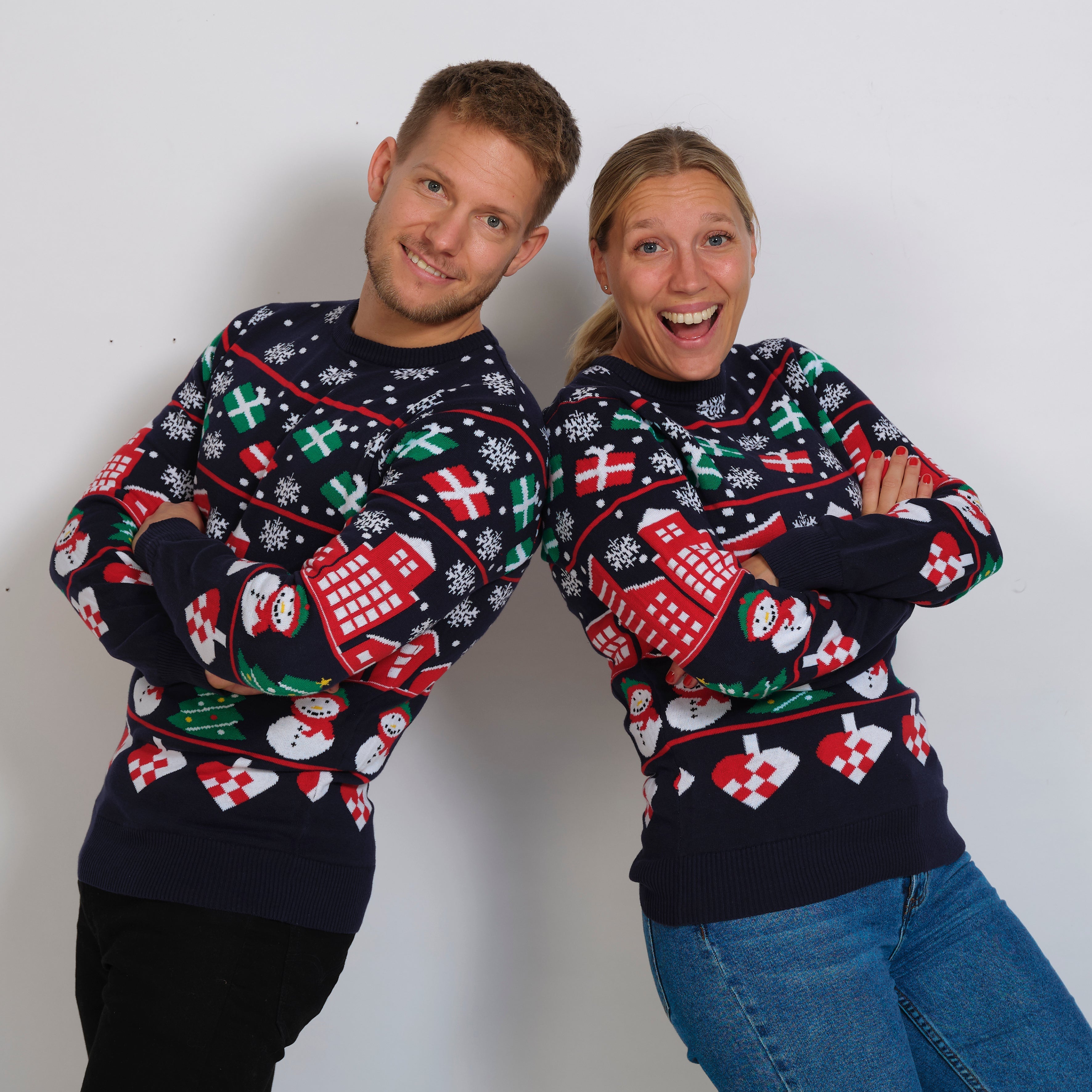 En dame og en mand, begge iført blå julesweatre med julehjerter, snemænd og gaver på.
